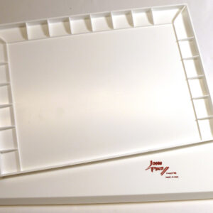 A large white rectangular plastic watercolor palette photographed at a slight angle from above. The palette tray features a spacious flat mixing area in the center surrounded on three sides by rows of square paint wells — approximately ten wells along the top edge and six wells along each side. The tray rests on a flat white rectangular lid slightly larger than the tray itself. A red handwritten "John Pike" signature logo with the words "Palette" and "Made in USA" is printed on the lower right of the lid.