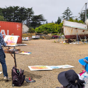 Watercolor artist standing in front of an easel which is distantly placed in front of an old ship in the yard for repair.
