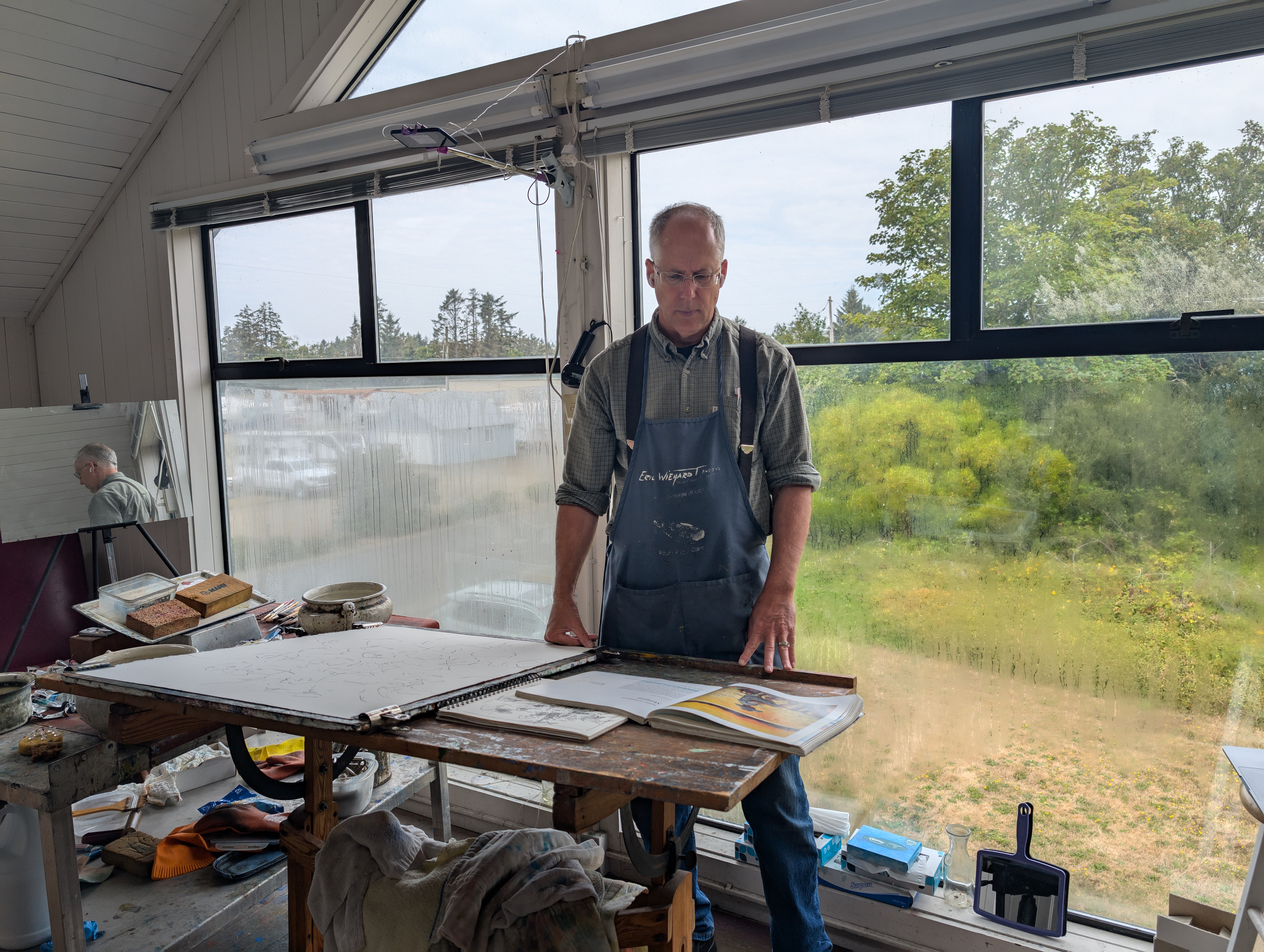 ric Wiegardt; Standing in his painting studio with easel in front and large window behind,