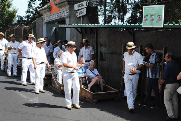 Several men in white clothing and straw hats are positioned around wicker toboggans on a paved road. Some toboggans are empty, while others contain seated passengers. Signage in the background indicates "CARREIROS DO MONTE" and directions to "FUNCHAL" and "BABOSAS".