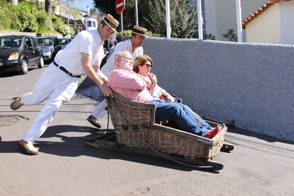 Two men in white uniforms and straw hats are propelling a wicker toboggan down a paved, sloping road. The toboggan carries a man in a pink shirt and a woman in sunglasses. Cars are parked along the side of the road, and a "Do Not Enter" sign is visible in the background.