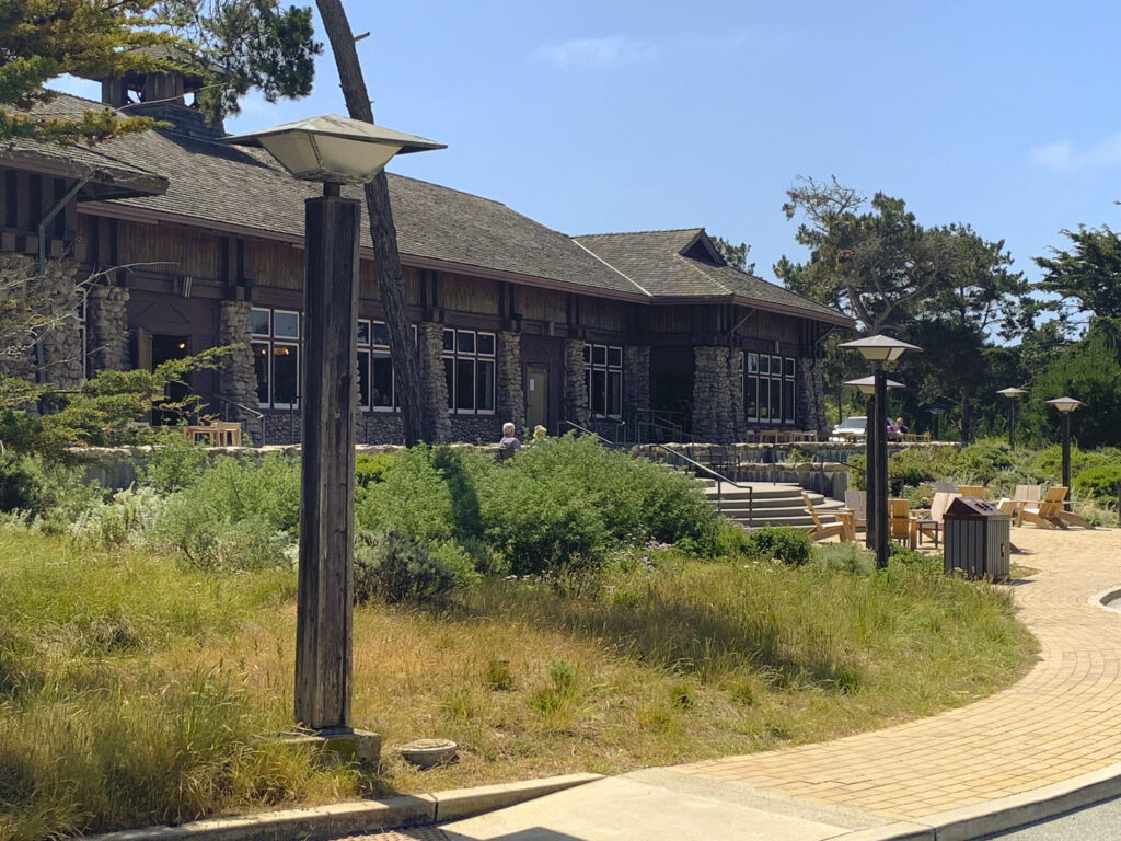 The rustic and elegant architecture of a large conference building at the Asilomar Conference Grounds under a clear blue sky. The building features a dark shingled roof, stone walls, and numerous windows, blending into the surrounding landscape. In the foreground, a paved pathway curves, flanked by natural, somewhat dry coastal grasses and low green shrubs. Several wooden outdoor chairs and lampposts are visible, suggesting a welcoming and open environment.