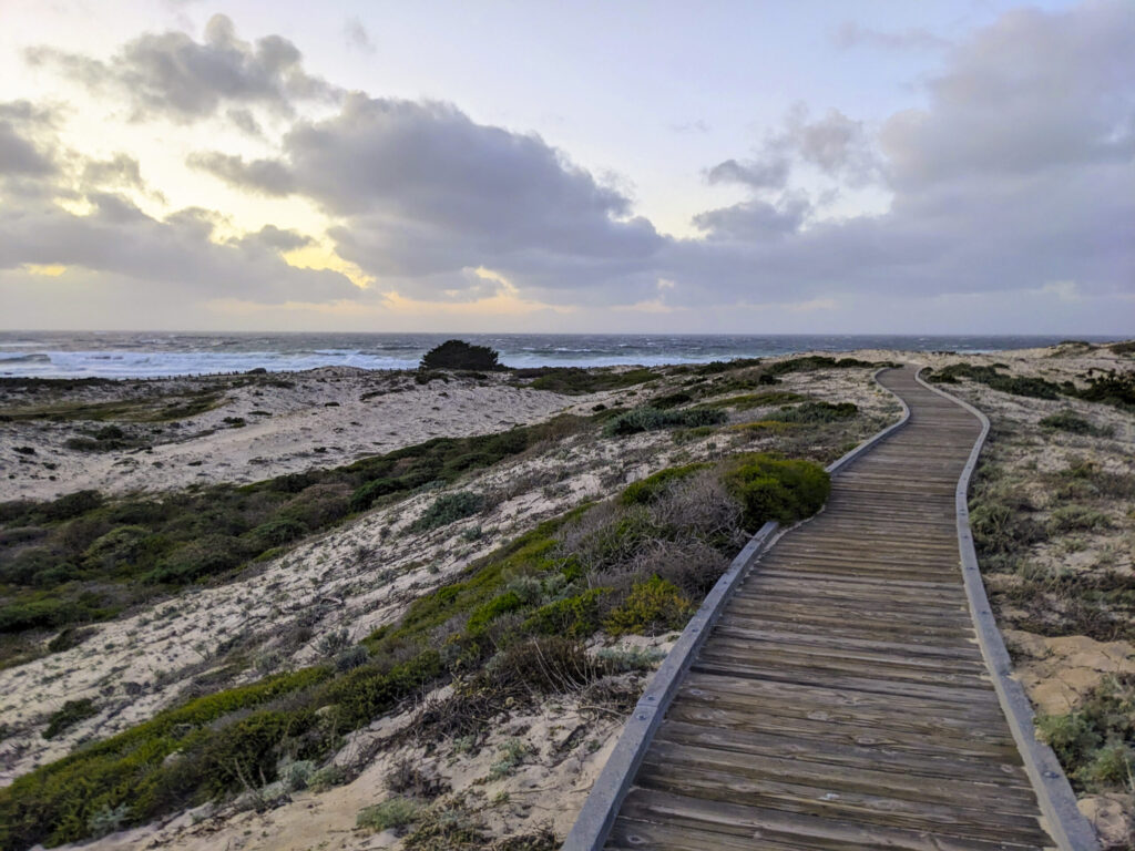 A winding wooden boardwalk stretches into the distance over sandy dunes covered with sparse, low-lying coastal vegetation. To the left, the rough ocean waves crash under a dramatic, overcast sky with hints of a distant sunset or sunrise glow. The scene conveys the rugged beauty of a coastal landscape at dusk or dawn.