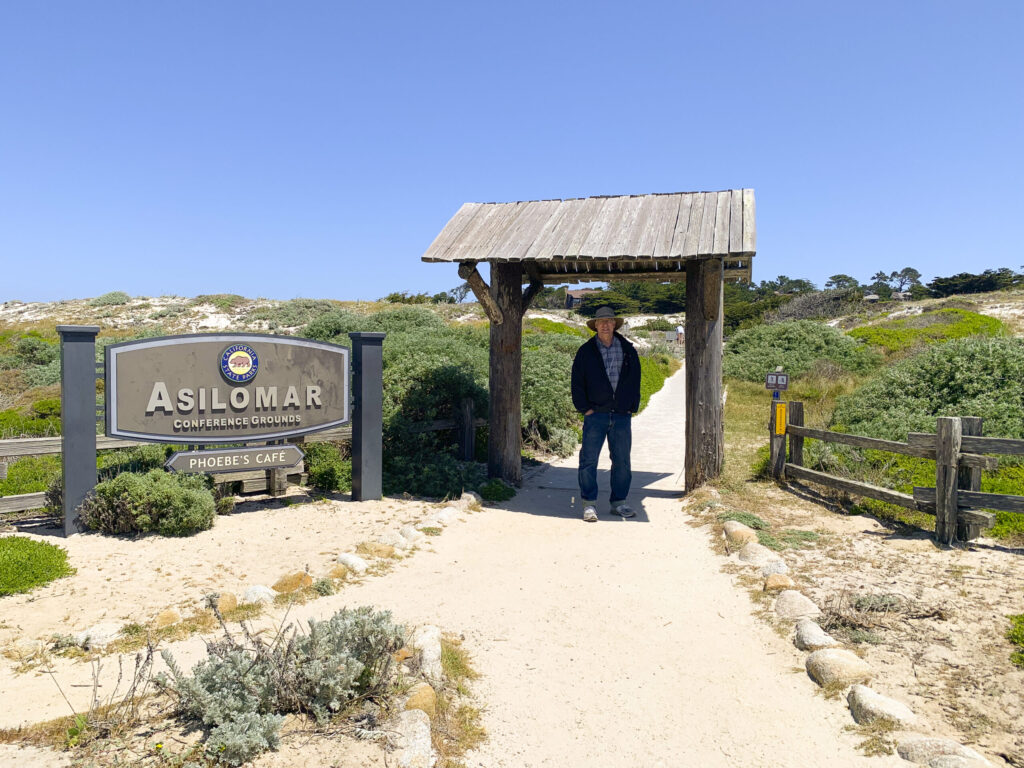 Watercolor artist Eric Wiegardt stands smiling on a sandy path, dressed in a dark jacket, blue jeans, and a wide-brimmed hat, beneath a rustic wooden archway. To his left, the 'Asilomar Conference Grounds' sign, a large grey sign with silver lettering and the California State Parks logo, stands prominently amidst coastal dunes and green foliage. The sky above is clear and blue, indicating a bright, sunny day at Asilomar.