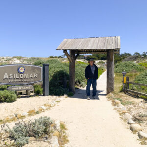 Watercolor artist Eric Wiegardt stands smiling on a sandy path, dressed in a dark jacket, blue jeans, and a wide-brimmed hat, beneath a rustic wooden archway. To his left, the 'Asilomar Conference Grounds' sign, a large grey sign with silver lettering and the California State Parks logo, stands prominently amidst coastal dunes and green foliage. The sky above is clear and blue, indicating a bright, sunny day at Asilomar.