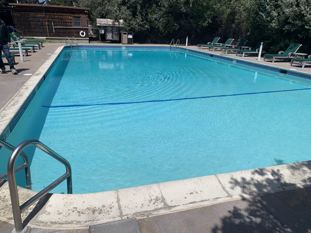 A clear, rectangular outdoor swimming pool with bright blue water and a dark blue lane line visible across its width. The pool is surrounded by a light-colored concrete deck. On the right, several green lounge chairs are arranged, and on the left, a metal handrail leads into the water. In the background, a rustic wooden building and lush green trees are visible under a bright, sunny sky.