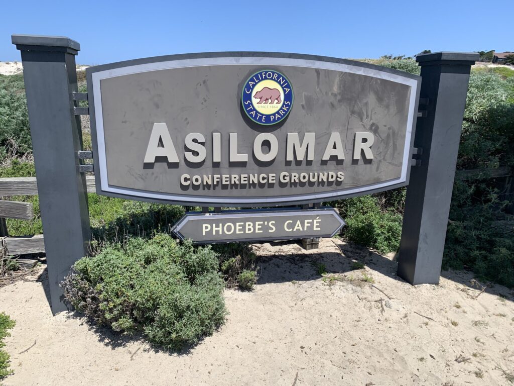 A large, prominent sign for 'Asilomar Conference Grounds' with 'California State Parks' logo at the top. The sign is grey with raised silver letters, set between two dark grey pillars. Below the main sign, a directional arrow points left towards 'Phoebe's Café.' The sign is surrounded by sandy ground and lush green coastal vegetation under a bright blue sky, welcoming visitors to the historic site.