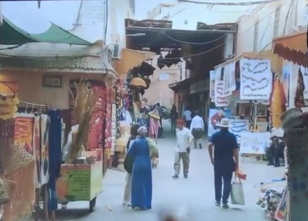 A photo of a bustling Moroccan market street, with stalls displaying goods, people walking, and awnings covering the narrow alleyway.