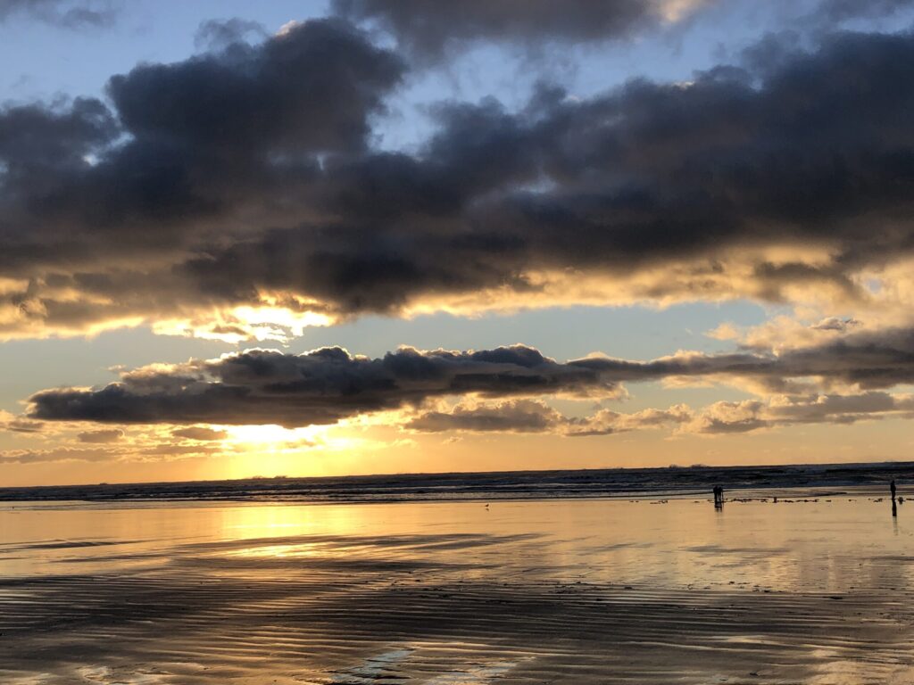 A stunning reference photo of a sunset over a beach, featuring a dramatic sky with heavy clouds framing the sun's golden light. The image provides an excellent study of light, color, and reflections on the wet, rippling sand.