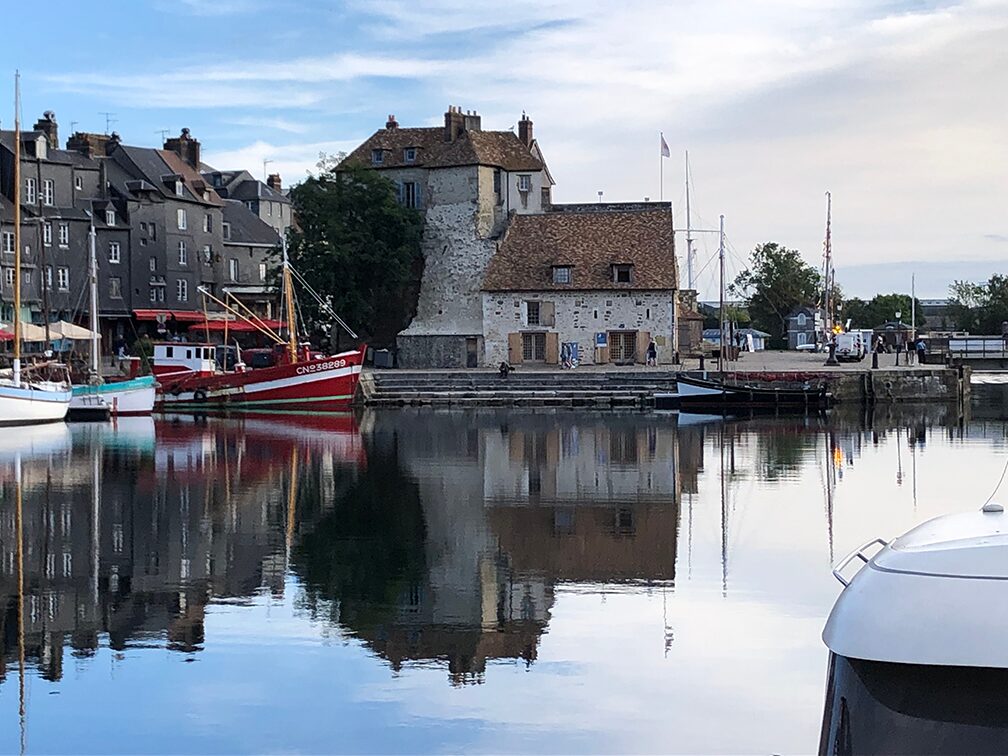 A view of a historic harbor with a prominent stone building at its center. The calm water reflects the building, surrounding old houses, and the docked boats, including a red fishing boat and several sailboats. The scene is set under a partly cloudy sky.