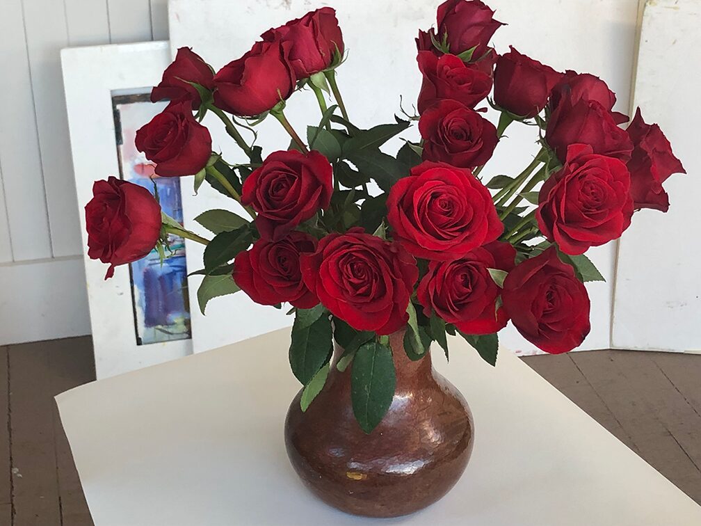 A large bouquet of bright red roses with green stems in a round, bronze-colored vase
