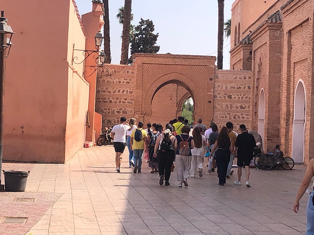 A group of people approaches a large arched entrance in a Moroccan alley, surrounded by tall walls with traditional brick patterns and muted tones. The scene reflects cultural exploration and architectural heritage.