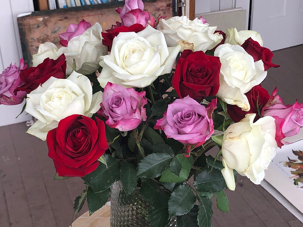 A close-up photograph of a vibrant bouquet of roses in a glass vase, featuring white, deep red, and mauve/pink roses with green leaves.