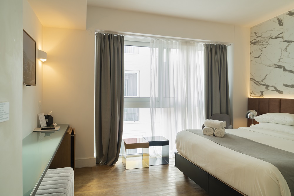 A photograph of a modern hotel room, showcasing a bright and clean aesthetic. The room features a large bed with a dark grey throw, a marble-patterned accent wall, and a large window with sheer and heavy curtains, suggesting a comfortable and well-lit space.