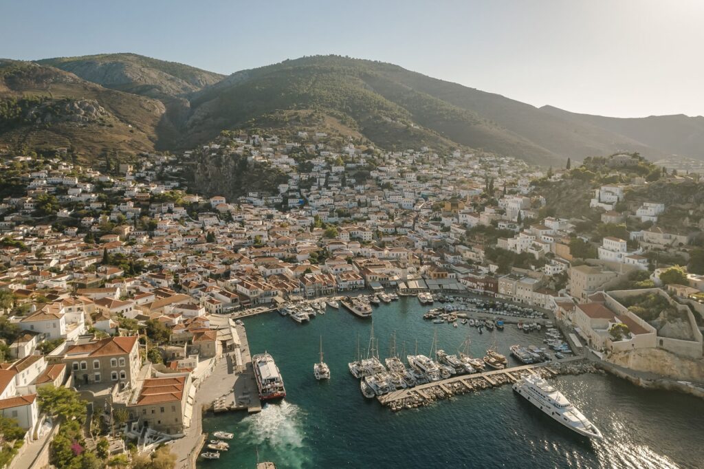  An aerial view of a picturesque coastal town built on a hillside, with a bustling harbor filled with boats and white and terracotta-roofed buildings.