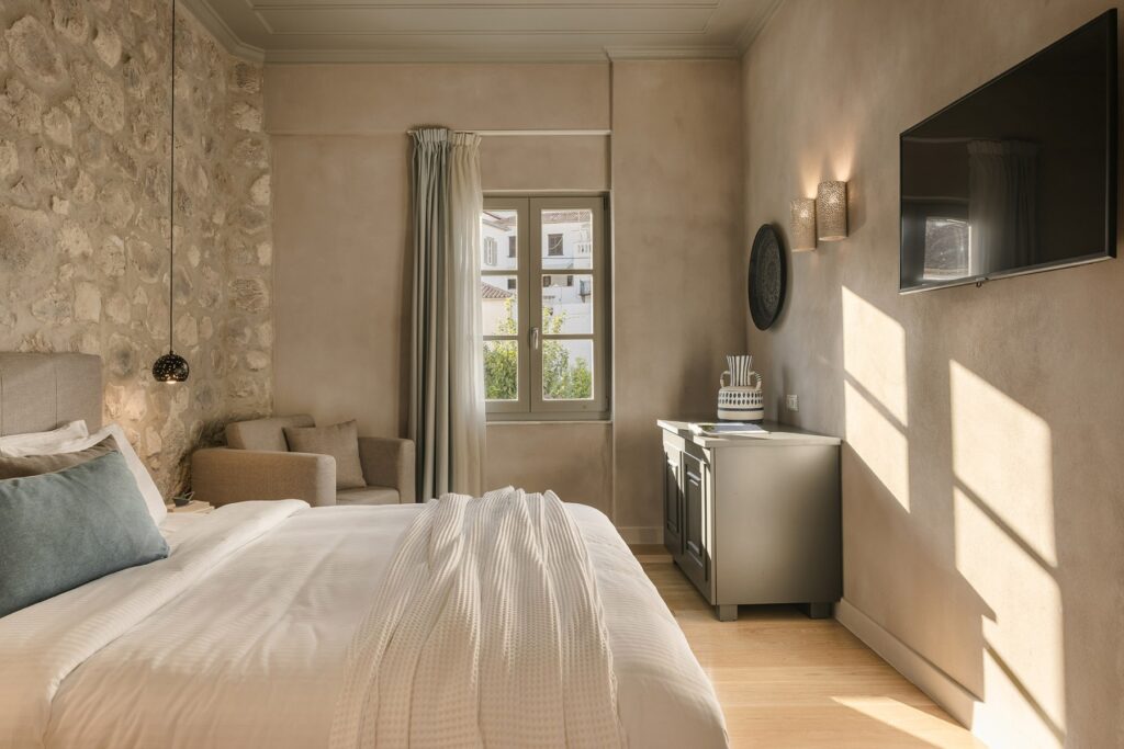 A photograph of a room at the Hydroussa Hotel, blending modern comforts with rustic charm. The room features a bed with a neutral duvet, a striking exposed stone wall, and a smooth, neutral-toned wall with a flat-screen TV. The small window and wooden floor complete the serene aesthetic.