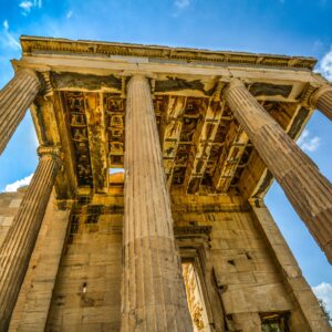 A low-angle photograph of the ancient Parthenon in Athens. The image highlights the massive, fluted columns and the weathered stone of the structure against a dramatic blue and cloudy sky.