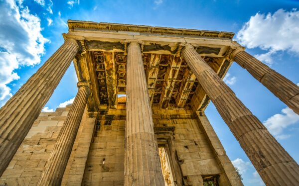 A low-angle photograph of the ancient Parthenon in Athens. The image highlights the massive, fluted columns and the weathered stone of the structure against a dramatic blue and cloudy sky.