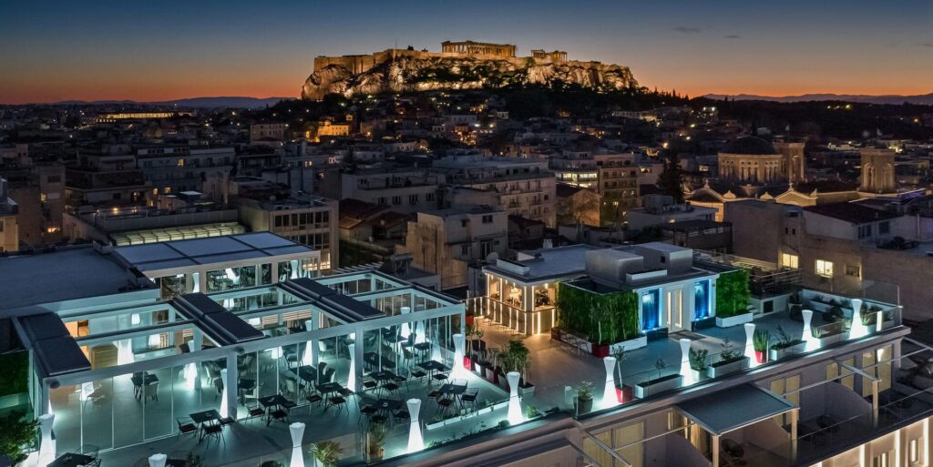 An evening view from a modern rooftop terrace, showing a lit outdoor seating area overlooking the city of Athens. The ancient Acropolis and Parthenon are prominently illuminated on a hill in the background against a twilight sky.