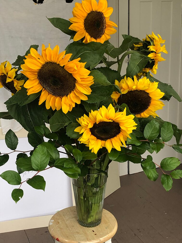 A vertical shot of a vibrant bouquet of sunflowers in a clear glass vase. The vase sits on a small, round wooden stool. The sunflowers have bright yellow petals and dark brown centers, surrounded by large, green leaves.