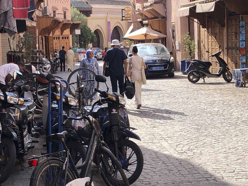 A busy, sunlit cobblestone street in Morocco with people walking past parked mopeds and bicycles. Buildings with arched doorways line the road.