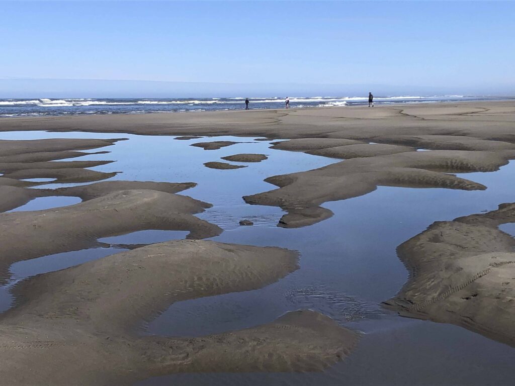 A wide, flat sandy beach at low tide with large blue-tinted tide pools reflecting a clear sky and tiny figures walking in the distance.