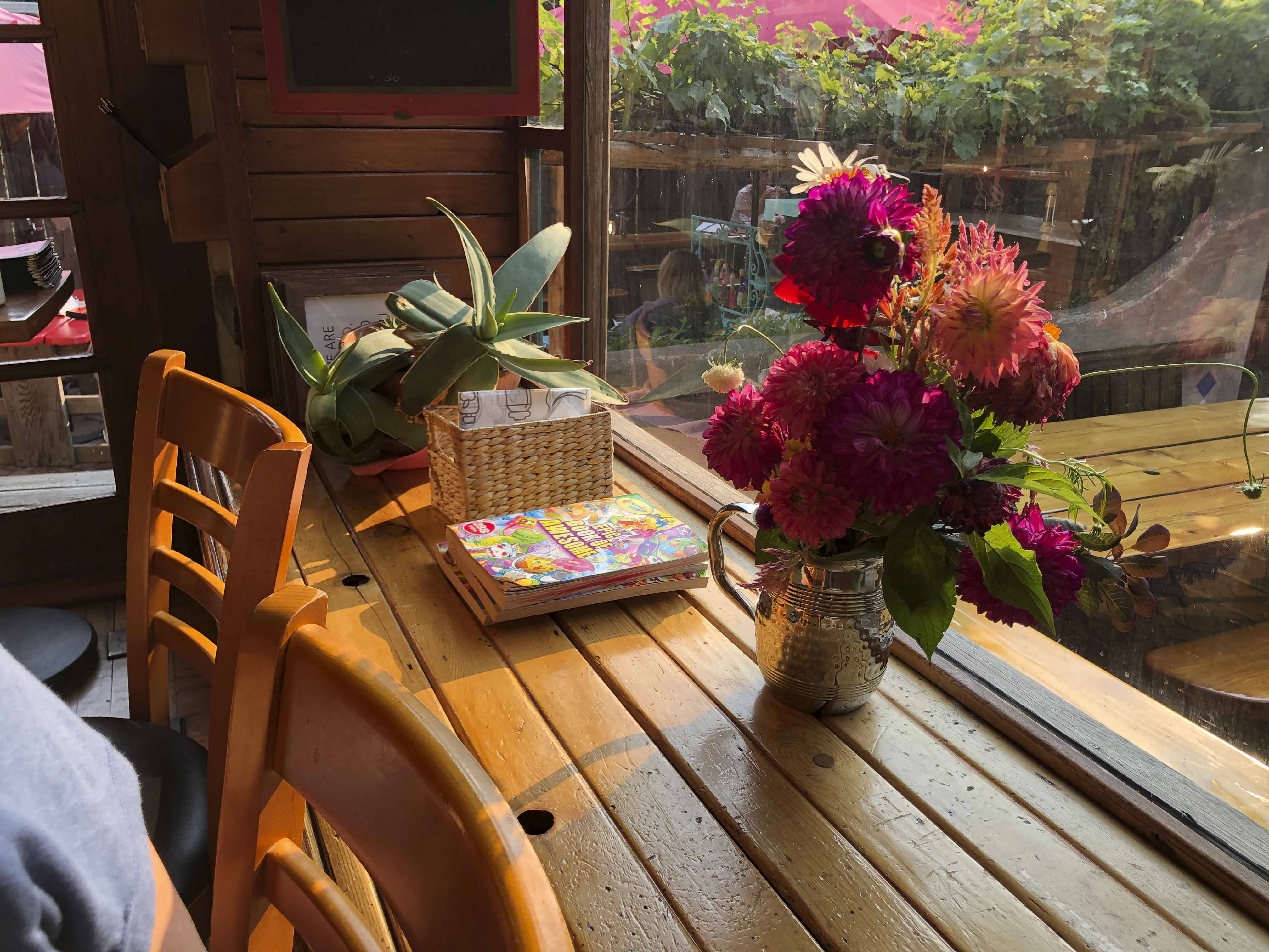 A sunlit still life on a wooden table near a window, featuring a vase of pink flowers, a potted aloe plant, and a stack of books.