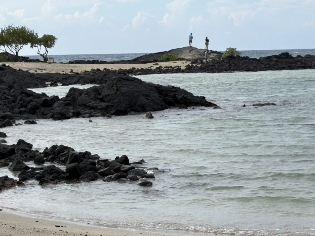 A landscape photograph of a rocky coastline. In the foreground, dark volcanic rocks meet a pale sandy beach and light blue water with gentle ripples. In the distance, three people stand atop a large rock formation under a pale, cloudy sky. A small cluster of green trees sits on the left.