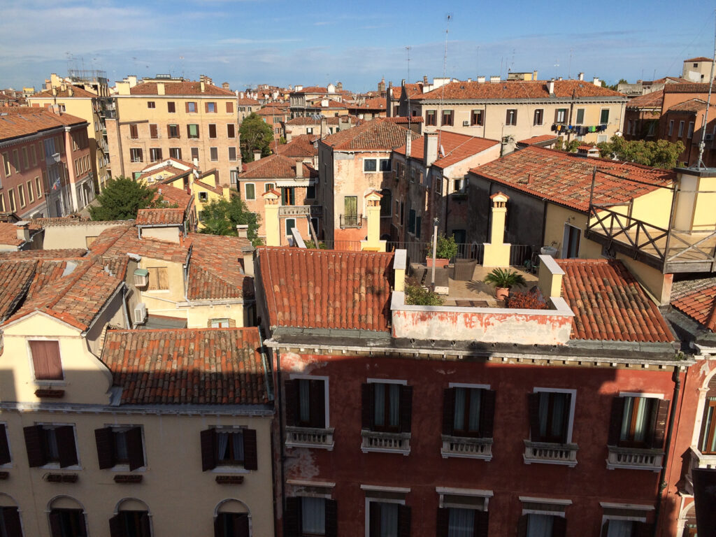 A wide-angle photograph looking over the terracotta tile rooftops of a European city, featuring warm-toned buildings with window shutters under a clear blue sky.