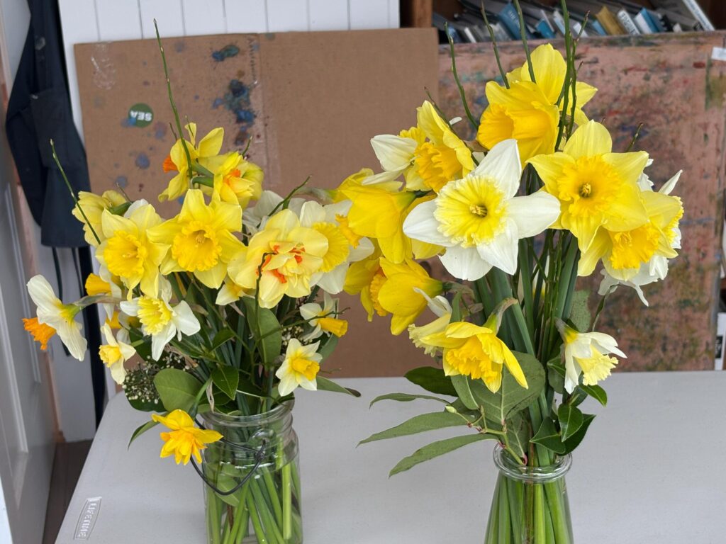 A photograph of two glass jars filled with bright yellow and white daffodils sitting on a white table in an artist's studio. The background shows a rustic workspace with a cardboard divider and various art supplies.