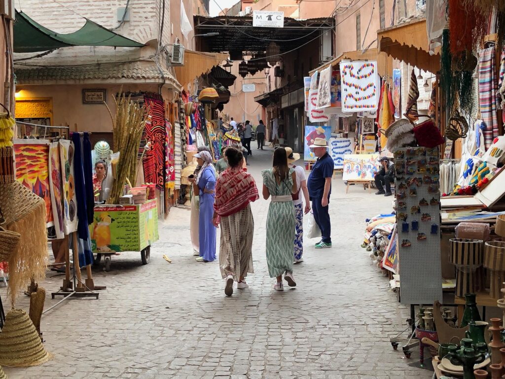 A reference photo of a Moroccan medina alleyway featuring shoppers walking past colorful textiles, rugs, and a cart of sugar cane. Warm terracotta walls and hanging tapestries create a narrow, sun-drenched corridor filled with texture.