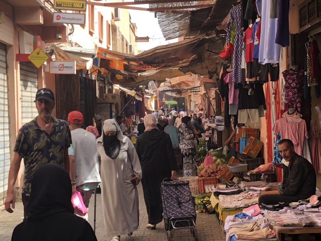 Warm golden light rakes through a narrow, awning-shaded Moroccan medina alley where shoppers in white djellabas and dark hijabs move toward a bright vanishing point, passing terracotta-toned walls, vivid green produce stalls, and colorful garments in red, blue, and pink hanging overhead.