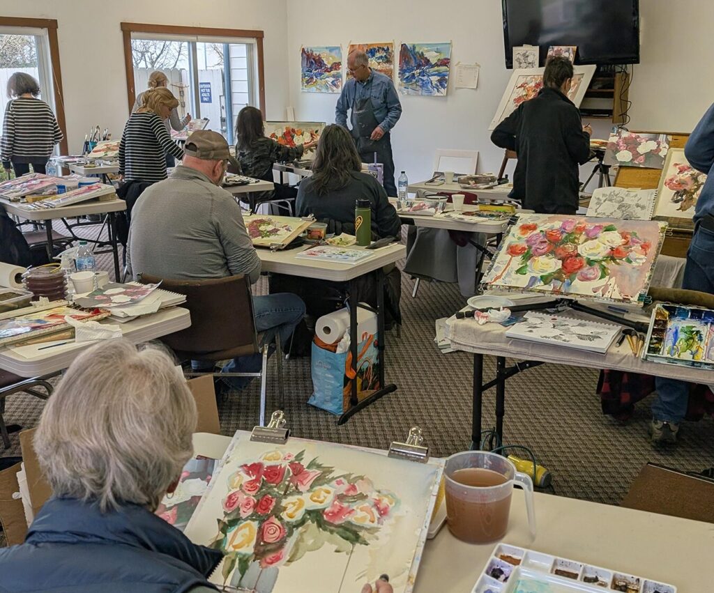 Students in an art studio paint colorful floral arrangements with watercolors while Eric Wiegardt, wearing a blue denim shirt and apron, provides instruction in the background. The scene is filled with white folding tables, vibrant red and yellow paintings, and large windows that let in soft, natural light.