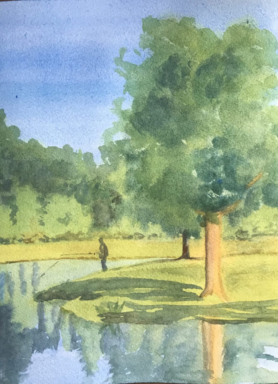 Late Summer Sun, original watercolor painting in warm tones of a man fishing in a lake from a grassy bank with green trees around. The sun is shining through the trees giving it a golden hue. The blue sky is reflecting in the lake.